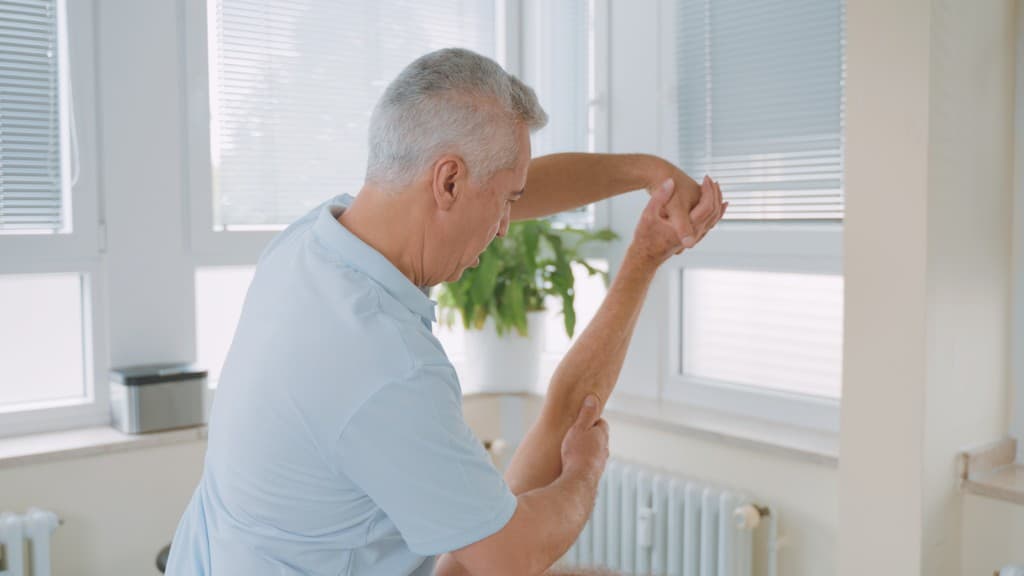 Walat Simo in der hellen Behandlungsraum-Praxis, sitzend auf der Therapieliege unter dem Schlingentisch-Gestell.